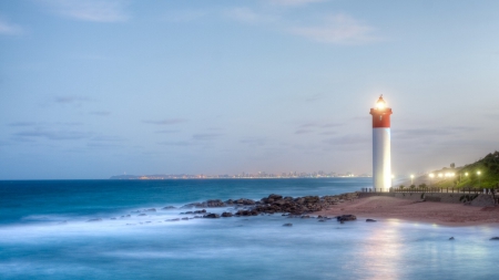 marvelous lit lighthouse with city in the background - lighthouse, lights, dusk, city, seashore