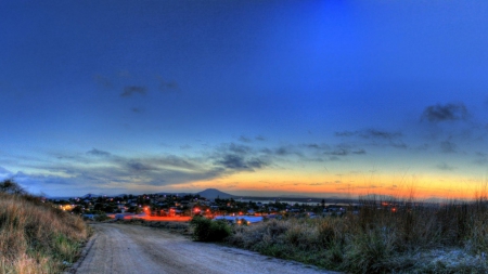 road down to a seaside town at dusk - dusk, town, lights, road, sea, weeds