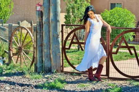 Cowgirl Style - posts, chain, window, stones, brunette, gate, cowgirl, building, skirt, yard, bushes, trees, wheel, gates, boots, wagon wheel