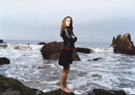 Eliza Duskku - standing, pleated black skirtr, ocean waves, brunette, black leather jacket, rocks