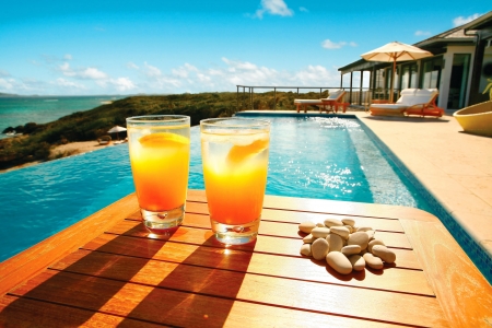 Two colds drinks and a pile of rocks sitting next to a beachside pool - sand, luxury, coast, tropical, rocks, orange, drinks, beach, shore, fancy, pool, swimming pool, water, drink, ocean